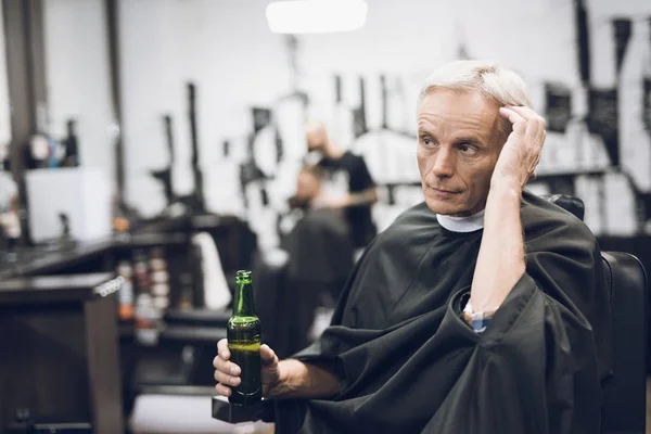 Den gamla mannen dricker alkohol i barberarens stol i barbershop. — Stockfoto