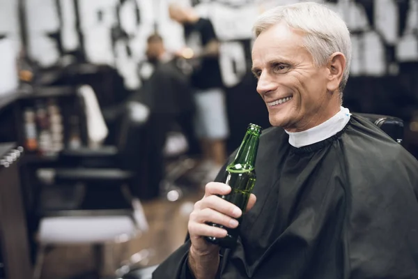De oude man drinkt alcohol in de Barbers stoel in de barbershop. — Stockfoto