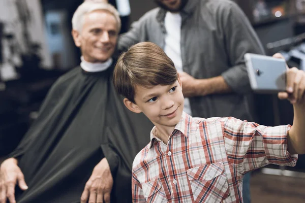 The boy makes selfie on a smartphone with two older men in barbershop.