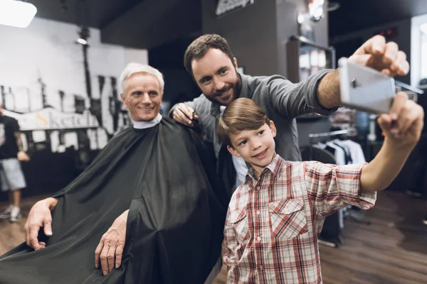 Le garçon fait du selfie sur un smartphone avec deux hommes plus âgés dans le salon de coiffure . — Photo