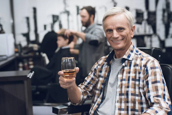 O velho bebe álcool na cadeira do barbeiro na barbearia. . — Fotografia de Stock