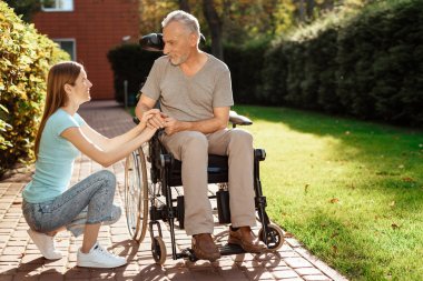 Yaşlı bir adam tekerlekli sandalyede oturuyor. Kız onun yanında çömelmiş, onu izliyor.