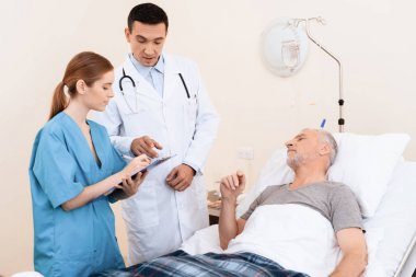Yaşlı adam tıbbi koğuşta yatak üzerinde yatıyor. Onun yanında bir doktor ve bir hemşire olduğunu.