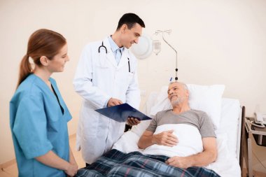 Yaşlı adam tıbbi koğuşta yatak üzerinde yatıyor. Onun yanında bir doktor ve bir hemşire olduğunu.