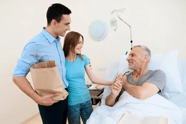 Um homem idoso jaz em um quarto de hospital em uma cama. Ele é visto por um homem com uma mulher. Estão ao lado do beliche dele. . — Fotografia de Stock