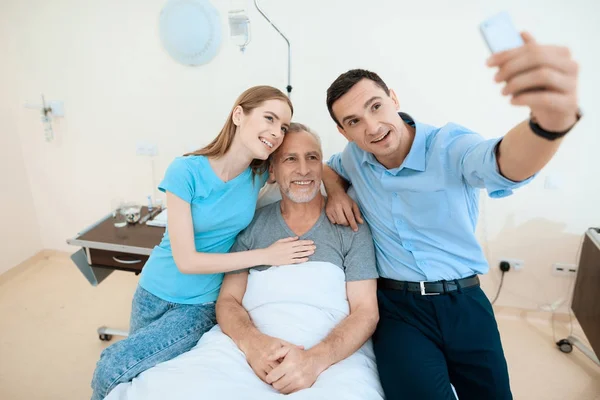 老人は、病室のベッドの上にあります。彼は、男と女で見られています。彼らはスマート フォンで selfie を行う. — ストック写真