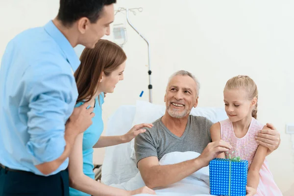 Um homem idoso jaz em um quarto de hospital em uma cama. Ele é visto por um homem com uma mulher e neta . — Fotografia de Stock