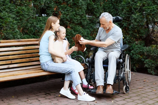 Um homem idoso está sentado em uma cadeira de rodas. Ele é visto por uma mulher com uma menina. Eles estão sentados em um banco no parque . — Fotografia de Stock