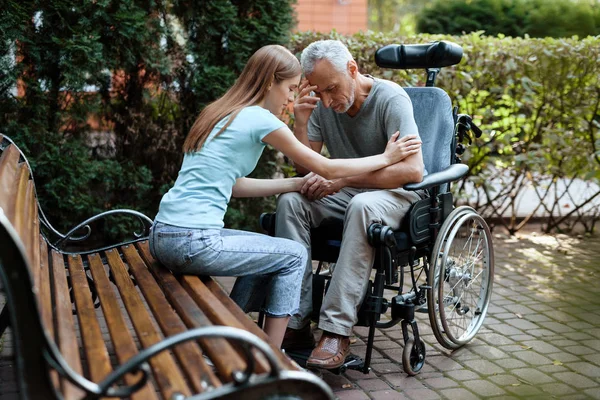 En äldre man sitter i rullstol. Bredvid bänken är hans dotter. De håller händerna. — Stockfoto