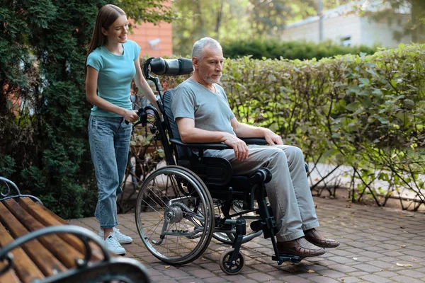 Um homem idoso está sentado em uma cadeira de rodas. Perto está sua filha, ela está pronta para levar o velho . — Fotografia de Stock