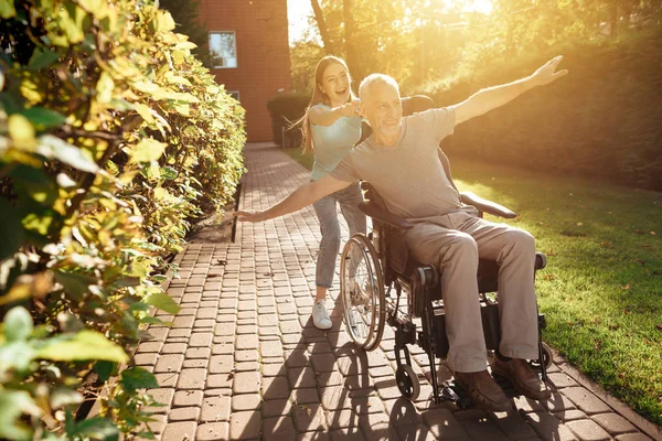 En äldre man sitter i rullstol. Flickan rullar det och de lura. De går utanför och skratta. — Stockfoto