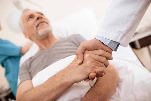 El anciano yace en una cuna en el pabellón médico, y junto a él se encuentra un médico. El doctor le da la mano al viejo . —  Fotos de Stock