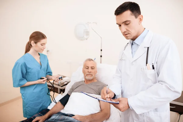 Gammal man lögner på en spjälsäng i medicinsk ward, och bredvid honom det är en läkare och en sjuksköterska. — Stockfoto