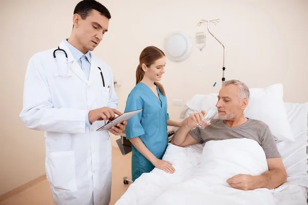 El anciano yace en una cuna en el pabellón médico, y junto a él hay un médico y una enfermera . —  Fotos de Stock