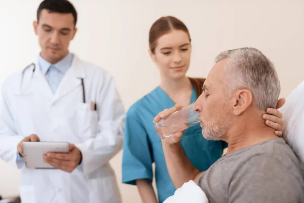 Gammal man lögner på en spjälsäng i medicinsk ward, och bredvid honom det är en läkare och en sjuksköterska. — Stockfoto