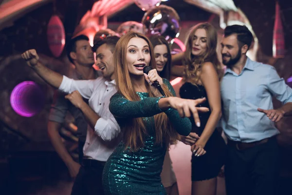 Una mujer con un vestido verde está cantando canciones con sus amigos en un club de karaoke. Sus amigos se divierten en el fondo . — Foto de Stock