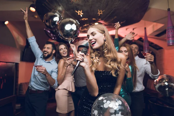Ungdomar har kul i en nattklubb och sjunga karaoke. I förgrunden finns det en kvinna i en svart klänning. — Stockfoto