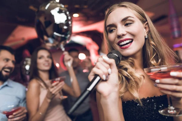 Young people have fun in a nightclub and sing in karaoke. In the foreground there is a woman in a black dress.