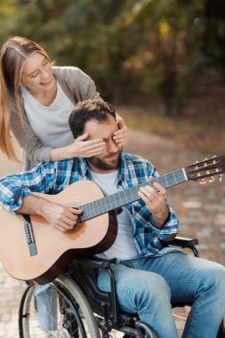Parkta gitar çalmak bir tekerlekli sandalye bir kişi. Bir kadın ona arkadan gelmiş ve elleriyle gözlerini kapalı.