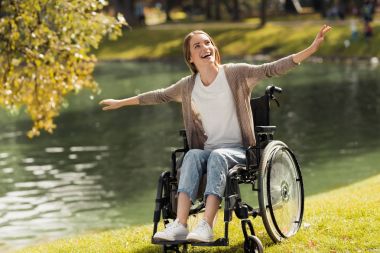 A woman in a wheelchair sits on the shore of a lake. She spread her arms out and laughed. clipart
