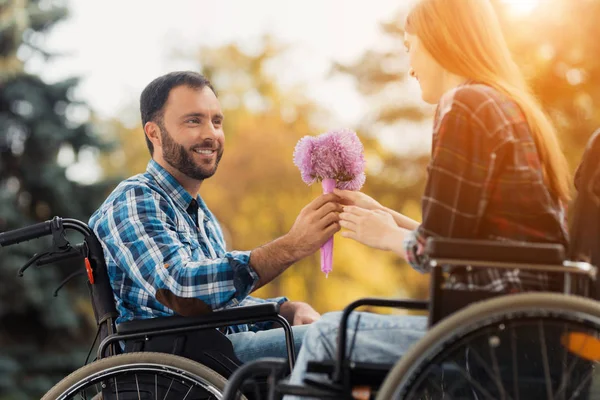 Uns inválidos em cadeiras de rodas encontraram-se no parque. Um homem está dando um buquê de flores para uma mulher . — Fotografia de Stock