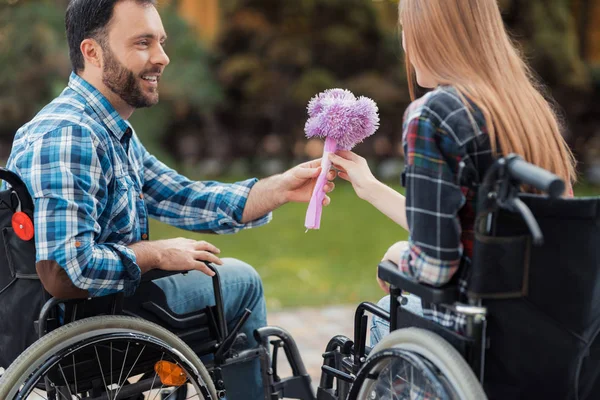 Uns inválidos em cadeiras de rodas encontraram-se no parque. Um homem está dando um buquê de flores para uma mulher . — Fotografia de Stock