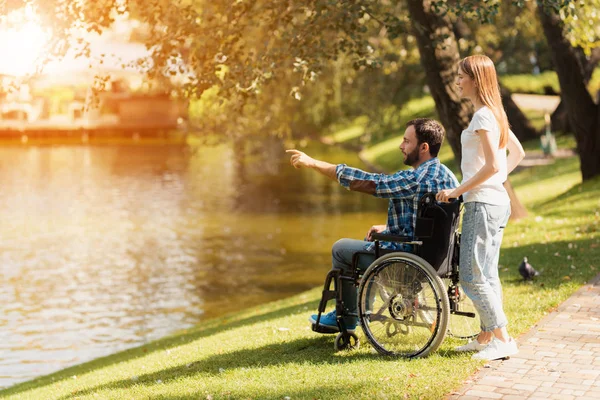 Una mujer está caminando en el parque con un hombre en silla de ruedas. Están en la orilla de un lago . — Foto de Stock