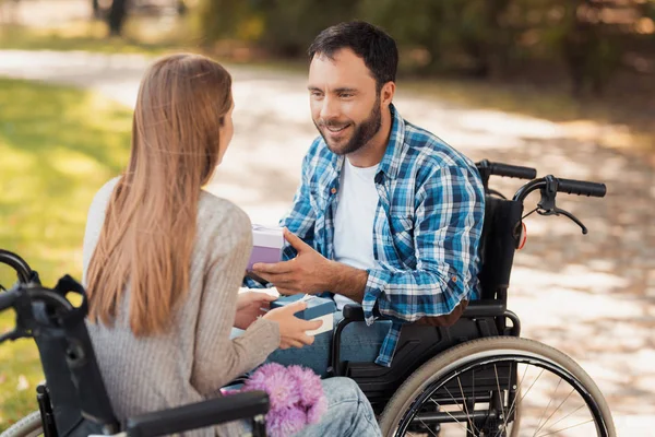 Un hombre y una mujer en silla de ruedas se conocieron en el parque. Se intercambian regalos y se sonríen. . — Foto de Stock