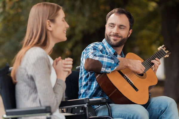 В парке встретились мужчина и женщина в инвалидных колясках. Мужчина играет на гитаре, женщина слушает и улыбается . — стоковое фото