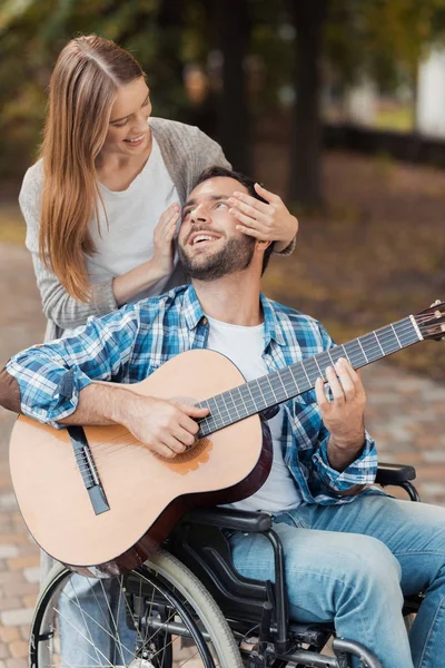一个坐在轮椅上的人在公园里弹吉他。一个女人从背后向他走来, 用她的双手闭上眼睛. — 图库照片