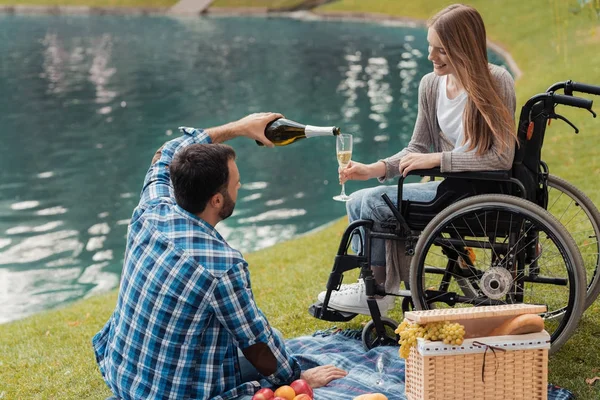 Homme et femme en fauteuil roulant dans le parc. Un homme s'assoit sur une couverture et verse à une femme une coupe de champagne dans un verre . — Photo