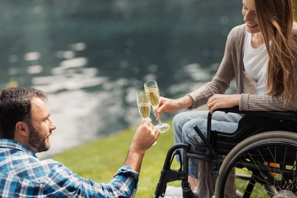 Frau im Rollstuhl im Park mit einem Mann. Ein Mann sitzt auf einer Decke. Sie klirren mit Champagner. — Stockfoto