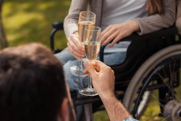 Закрывай. Женщина в инвалидном кресле в парке с мужчиной. Мужчина сидит на одеяле. Они звонят в бокалы с шампанским . — стоковое фото