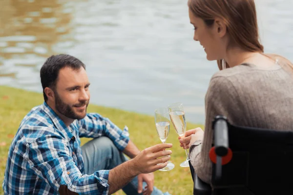 女人坐在轮椅上和一个男人在公园里。一个人坐在毯子上。他们用香槟碰杯. — 图库照片