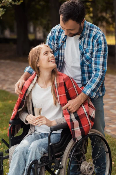 女人坐在轮椅上和一个男人在公园里。男人用格子格子布遮住肩膀。. — 图库照片