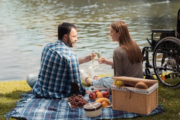 一个男人和一个女人坐在湖岸边的地毯上野餐。附近有一个轮椅. — 图库照片
