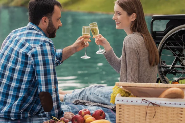 En man och en kvinna sitter vid sjön på en matta för picknick. I närheten finns är en rullstol. — Stockfoto