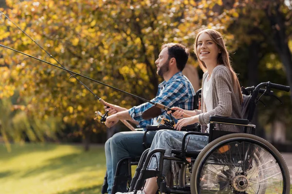 Una mujer y un hombre en silla de ruedas están sentados en la orilla del lago. Tienen hilanderos en sus manos y están pescando. . — Foto de Stock