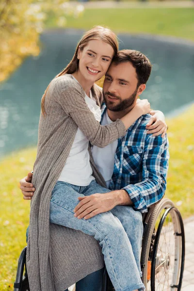 Um homem está sentado em uma cadeira de rodas em um belo parque verde. Uma mulher está sentada no colo dele e abraçando-o . — Fotografia de Stock