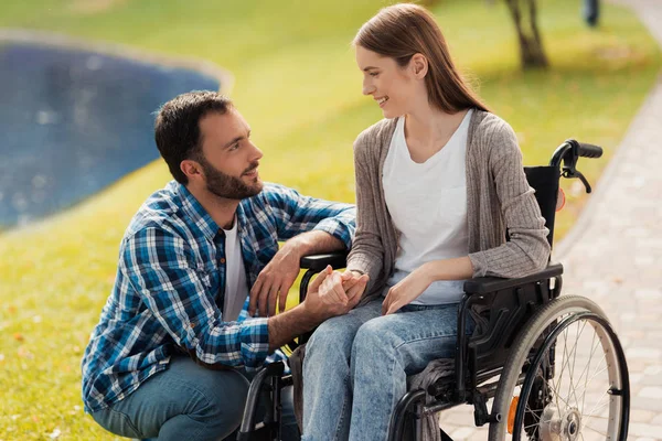 Una mujer está sentada en silla de ruedas. Un hombre está sentado a su lado y sosteniendo sus manos . —  Fotos de Stock