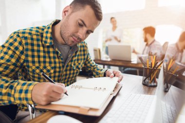 Tekerlekli sandalyede bir adam bir defter bir kalem ile yazar. O parlak bir ofiste çalışıyor.