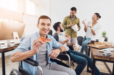 Tekerlekli sandalyede bir adam bir parça pizza tutan kameraya poz veriyor..