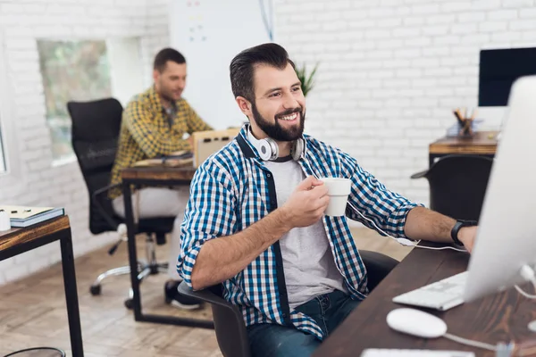 Un hombre está sentado en una oficina moderna y luminosa en la computadora, sosteniendo una taza de café o té en su mano . — Foto de Stock