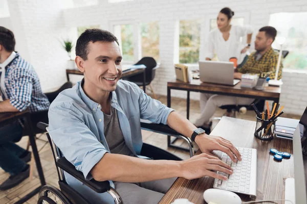 La persona con discapacidad en una silla de ruedas trabaja en el teclado de la computadora en una mesa en una oficina moderna y luminosa . — Foto de Stock