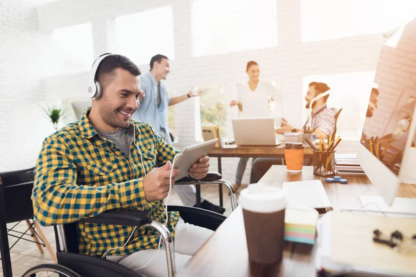 Чоловік у інвалідному візку працює з планшетом у сучасному офісі. Він слухає музику на навушниках . — стокове фото