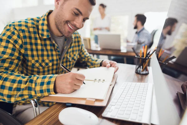 Schreibt ein Mann im Rollstuhl mit einem Stift in ein Notizbuch. er arbeitet in einem hellen Büro. — Stockfoto