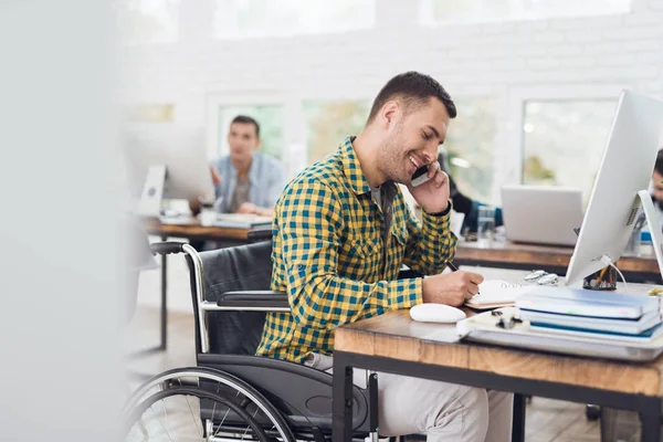Ένας άνθρωπος σε μια αναπηρική καρέκλα γράφει με ένα στυλό σε ένα σημειωματάριο και μιλάει στο τηλέφωνο. Εργάζεται σε ένα φωτεινό γραφείο. — Φωτογραφία Αρχείου