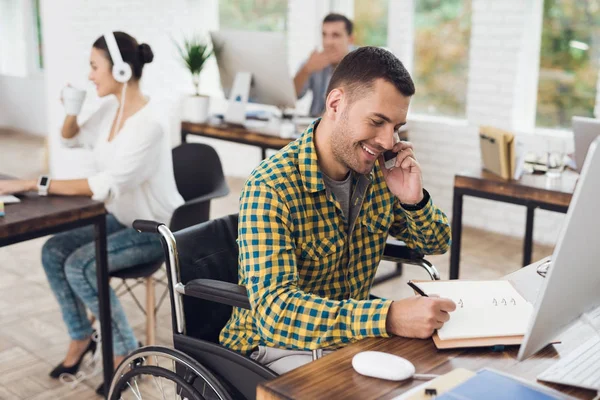 Un uomo su una sedia a rotelle scrive con una penna in un quaderno e parla al telefono. Lavora in un ufficio luminoso. . — Foto Stock