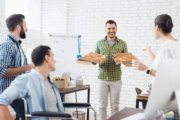 Trabalhadores de escritório e homem em uma cadeira de rodas estão comendo pizza. Eles trabalham em um escritório brilhante e moderno . — Fotografia de Stock