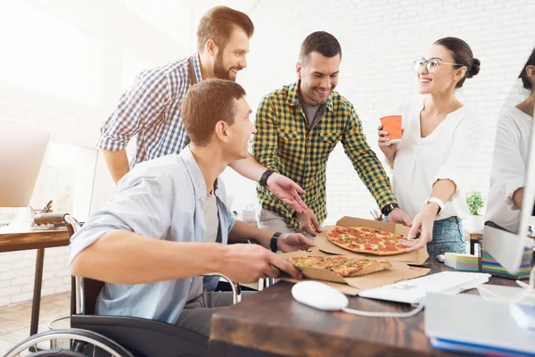 Trabalhadores de escritório e homem em uma cadeira de rodas estão comendo pizza. Eles trabalham em um escritório brilhante e moderno . — Fotografia de Stock